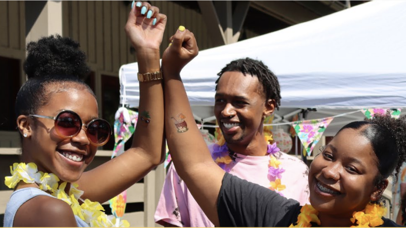 Three African-American people smile and laugh together, two of them placing their arms together.